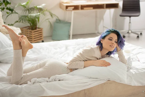 Belle femme aux cheveux colorés souriant et reposant avec les jambes croisées sur le lit — Photo de stock