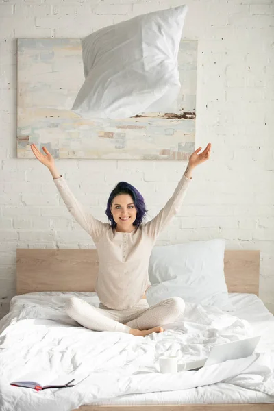 Freelancer mit bunten Haaren und überkreuzten Beinen wirft Kissen hoch, lächelt und blickt in die Kamera auf dem Bett — Stockfoto