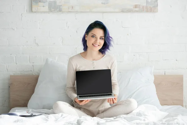 Freelance avec des cheveux colorés et jambes croisées montrant ordinateur portable, souriant et regardant la caméra sur le lit dans la chambre — Photo de stock