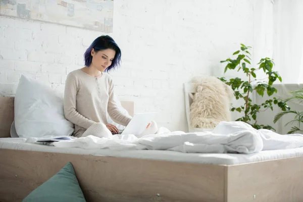 Freelancer com cabelos coloridos e pernas cruzadas trabalhando com laptop e sorrindo na cama no quarto — Fotografia de Stock