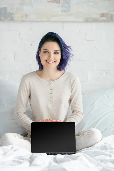 Freelance avec des cheveux colorés et jambes croisées montrant ordinateur portable, souriant et regardant la caméra sur le lit dans la chambre — Photo de stock