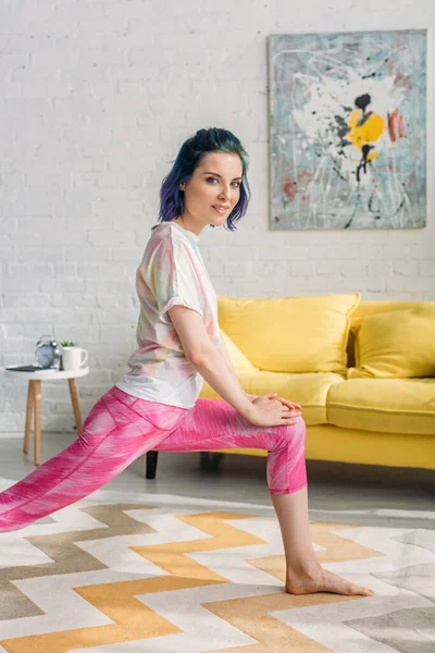 Menina com cabelo colorido fazendo ioga asana e olhando para a câmera na sala de estar — Fotografia de Stock