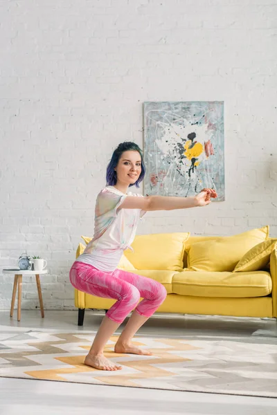 Chica con el pelo colorido en la silla posar con las manos extendidas, sonriendo y mirando a la cámara en la sala de estar - foto de stock