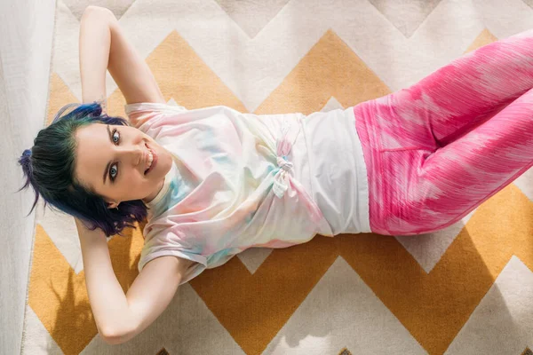 Top view of girl with colorful hair doing abs, looking at camera and smiling on floor in living room — Stock Photo