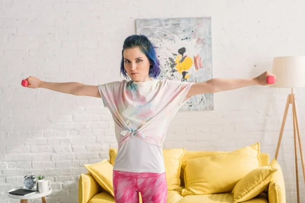 Woman with colorful hair holding dumbbells with outstretched hands and looking at camera in living room — Stock Photo