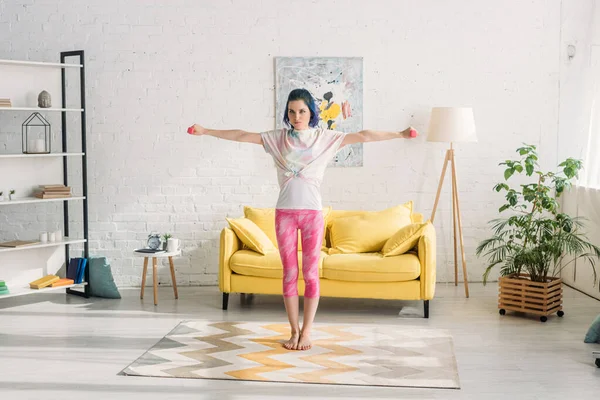 Konzentrierte Frau mit buntem Haar und ausgestreckten Händen, die Hanteln im Wohnzimmer halten — Stockfoto