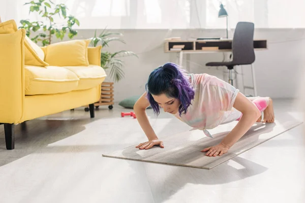 Chica con el pelo colorido haciendo flexiones en la alfombra de fitness en la sala de estar - foto de stock