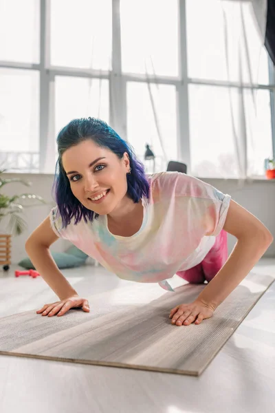 Menina com o cabelo colorido sorrindo, olhando para a câmera e fazendo flexões no tapete de fitness na sala de estar — Fotografia de Stock