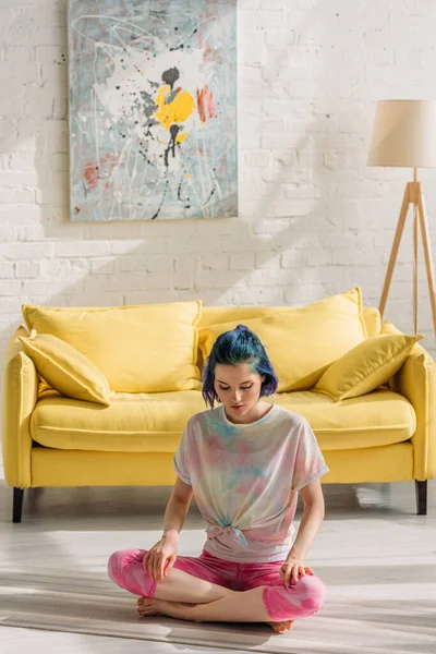 Ragazza con i capelli colorati e gambe incrociate seduta su tappetino yoga e meditando in soggiorno — Foto stock