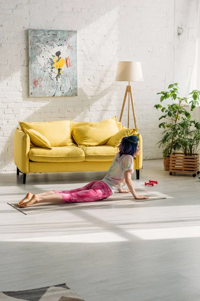 Fille aux cheveux colorés en pose de cobra sur tapis de yoga dans le salon — Photo de stock