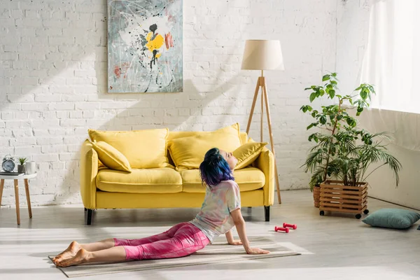 Девушка с красочными волосами в позу собаки вверх ногами на коврике для йоги в гостиной — стоковое фото
