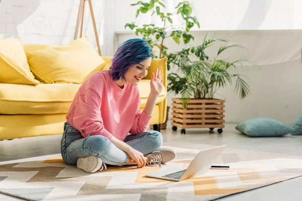 Chica con el pelo colorido agitando la mano, sonriendo y haciendo videollamada en el suelo en la sala de estar - foto de stock