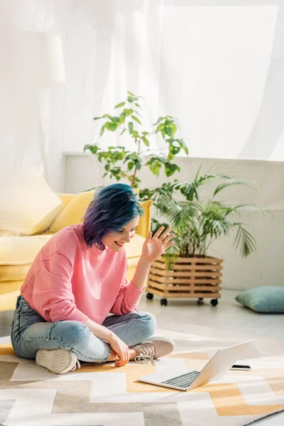 Chica con el pelo colorido agitando la mano, sonriendo y haciendo videollamada en el suelo - foto de stock