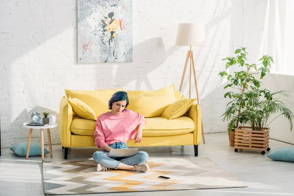 Freelancer with colorful hair and crossed legs working with laptop near sofa on floor — Stock Photo
