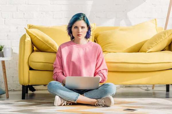 Freelance aux cheveux colorés et jambes croisées regardant la caméra et travaillant avec un ordinateur portable près du canapé dans le salon — Photo de stock