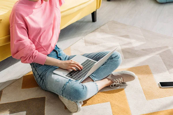Cropped view of freelancer with crossed legs working with laptop near sofa and smartphone on floor in living room — Stock Photo