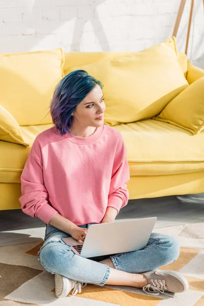 Nachdenkliche Freiberuflerin mit buntem Haar, die wegschaut und mit Laptop in der Nähe von Sofa auf dem Boden im Wohnzimmer arbeitet — Stockfoto
