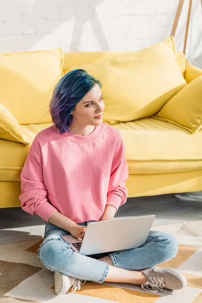 Pensiero freelance con i capelli colorati guardando altrove e lavorando con il computer portatile vicino divano al piano in soggiorno — Foto stock