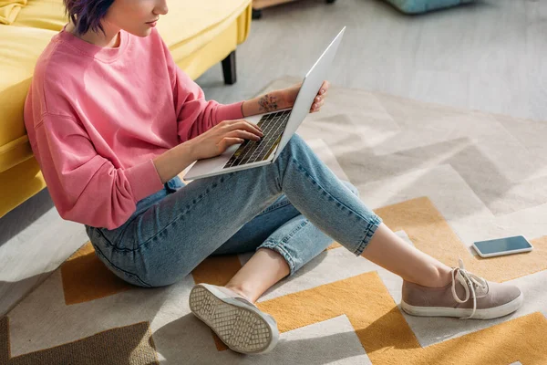 Vista recortada de freelancer com cabelo colorido trabalhando com laptop perto de sofá e smartphone no chão na sala de estar — Fotografia de Stock