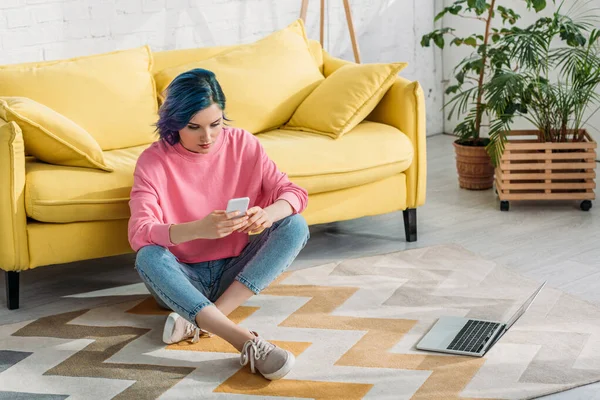 Freelancer with colorful hair and crossed legs holding smartphone on floor near sofa and laptop — Stock Photo