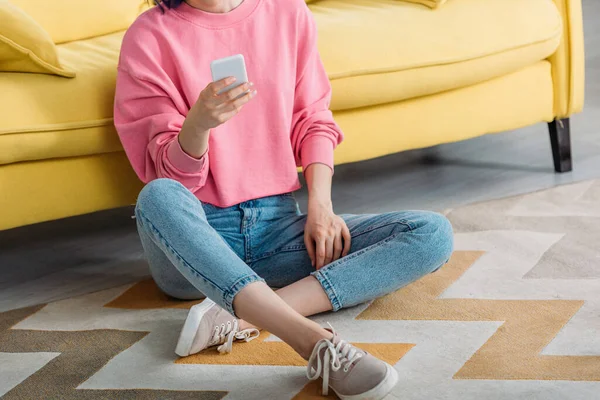 Vista cortada de mulher com pernas cruzadas segurando smartphone perto do sofá no chão — Fotografia de Stock