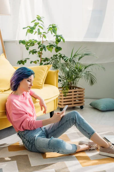 Mujer concentrada con cabello colorido sosteniendo teléfono inteligente cerca del sofá en el suelo en la sala de estar - foto de stock