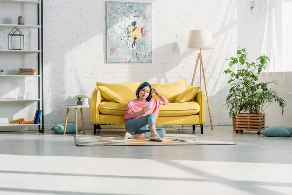 Femme avec des cheveux colorés et smartphone assis près du canapé sur le sol dans le salon — Photo de stock