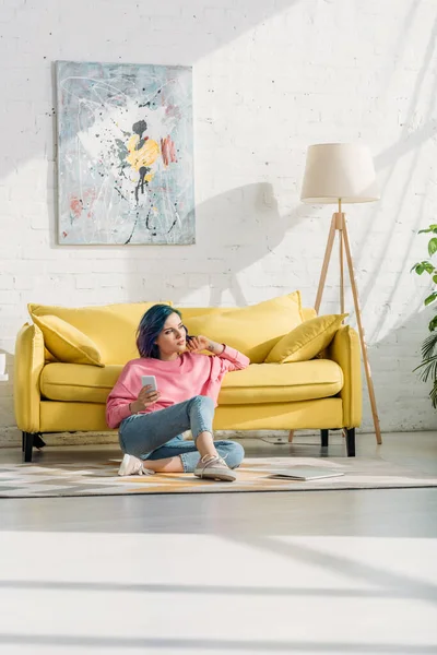 Femme réfléchie avec des cheveux colorés et smartphone assis près du canapé sur le sol dans le salon — Photo de stock