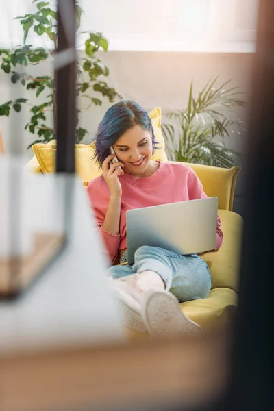 Foco seletivo de freelancer com cabelo colorido sorrindo e falando em smartphone com laptop no sofá — Fotografia de Stock