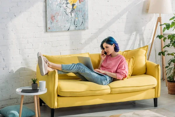 Фрилансер с красочными волосами разговаривает на смартфоне с ноутбуком и лежит на диване — стоковое фото