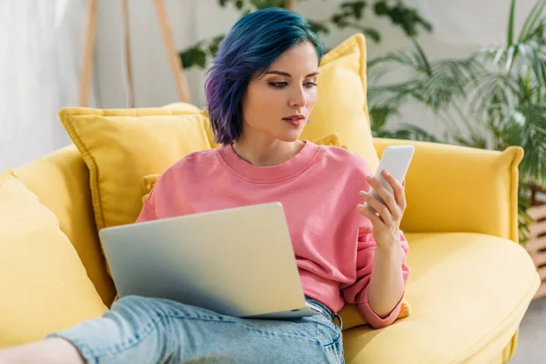 Freelancer with colorful hair and laptop looking at smartphone and lying on sofa — Stock Photo