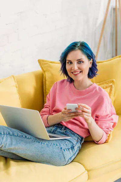 Freelance avec cheveux colorés, smartphone et ordinateur portable souriant et couché sur le canapé — Photo de stock