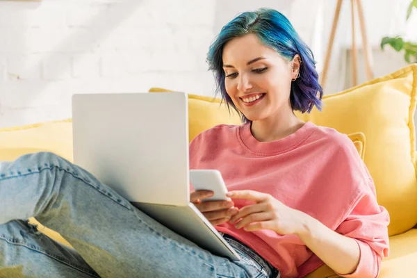Freelancer com cabelo colorido e laptop segurando smartphone, sorrindo e deitado no sofá — Fotografia de Stock