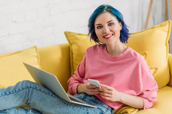 Freelancer com cabelo colorido e laptop segurando smartphone, sorrindo, olhando para a câmera e deitado no sofá — Fotografia de Stock