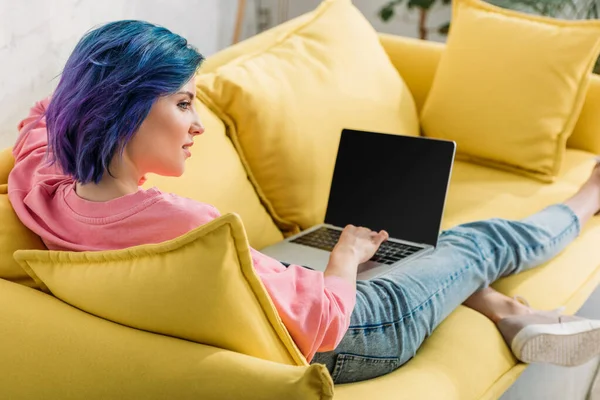 Фрилансер с раскрашенными волосами и ноутбуком улыбается и лежит на диване в гостиной — стоковое фото