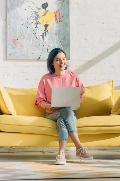 Freelancer con cabello colorido y portátil sonriendo y sentado en un sofá con las piernas cruzadas en la sala de estar - foto de stock