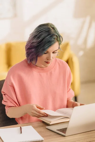 Freelancer with colorful hair reading book near laptop and copybook at table in living room — Stock Photo