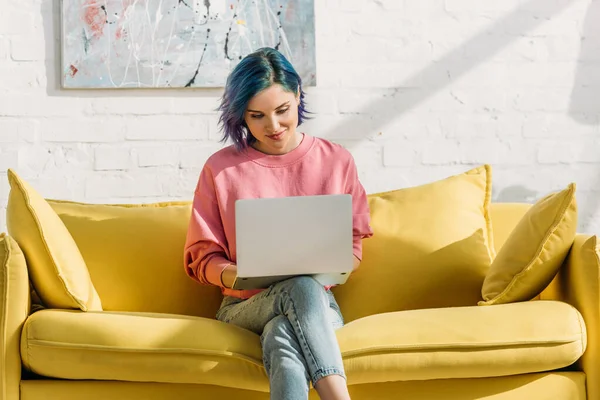 Freelancer with colorful hair and laptop smiling and sitting on yellow sofa with crossed legs in living room — Stock Photo