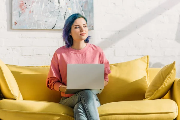 Freelancer com cabelo colorido e laptop olhando para longe e sentado no sofá amarelo com pernas cruzadas na sala de estar — Fotografia de Stock