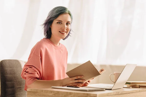 Freelancer mit buntem Haar und Buch blickt in die Kamera und lächelt am Tisch mit Laptop im Wohnzimmer — Stockfoto