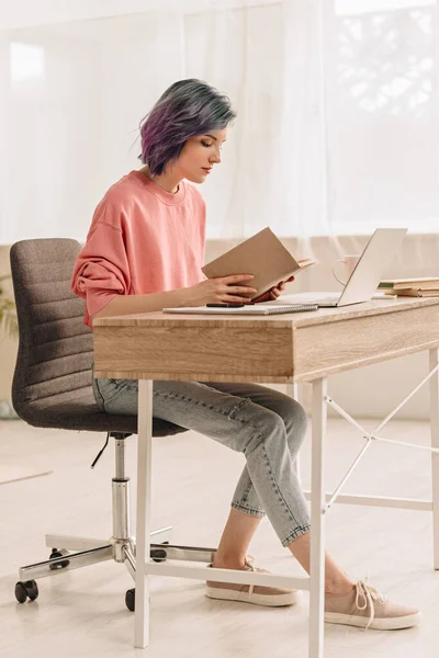 Freelancer com livro de leitura de cabelo colorido à mesa com laptop na sala de estar — Fotografia de Stock