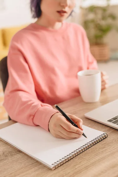 Vista ritagliata di freelance in possesso di tazza di tè e scrittura in notebook a tavola — Foto stock