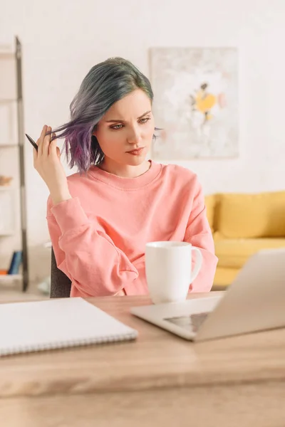 Foco seletivo de freelancer pensativo com caneta colorida cabelo segurando à mesa com laptop, xícara de chá e notebook na sala de estar — Fotografia de Stock