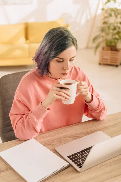 Vue grand angle de pigiste avec des cheveux colorés tenant tasse de thé à table avec ordinateur portable et ordinateur portable dans le salon — Photo de stock