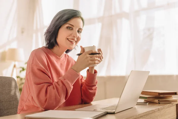 Freelancer mit buntem Haar, Tasse Tee und Stift blickt in die Kamera und lächelt am Tisch mit Laptop im Wohnzimmer — Stockfoto