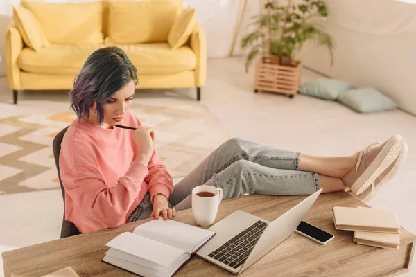Vista de alto ângulo do freelancer com cabelo colorido e caneta olhando para notebook e colocando pernas na mesa — Fotografia de Stock