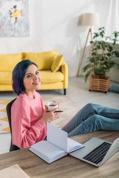 Vista ad alto angolo di freelance con capelli colorati, tazza di tè e penna sorridente e mettendo le gambe sul tavolo in soggiorno — Foto stock