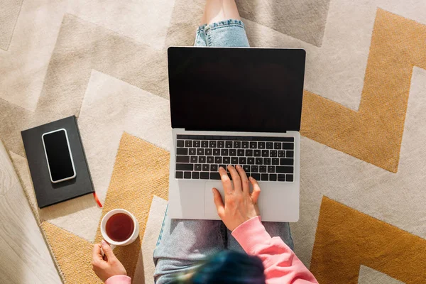 Vista cortada de freelancer com xícara de chá trabalhando com laptop perto de notebook e smartphone no chão na sala de estar — Fotografia de Stock