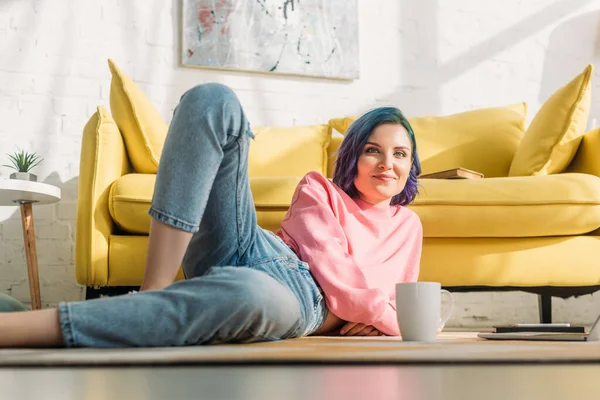 Selektiver Fokus einer Frau mit buntem Haar, die lächelt, in die Kamera blickt und auf dem Boden neben dem Sofa mit einer Tasse Tee im Wohnzimmer liegt — Stockfoto