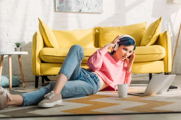 Freelancer com cabelos coloridos e fones de ouvido olhando para laptop e deitado perto de sofá e xícara de chá na sala de estar — Fotografia de Stock